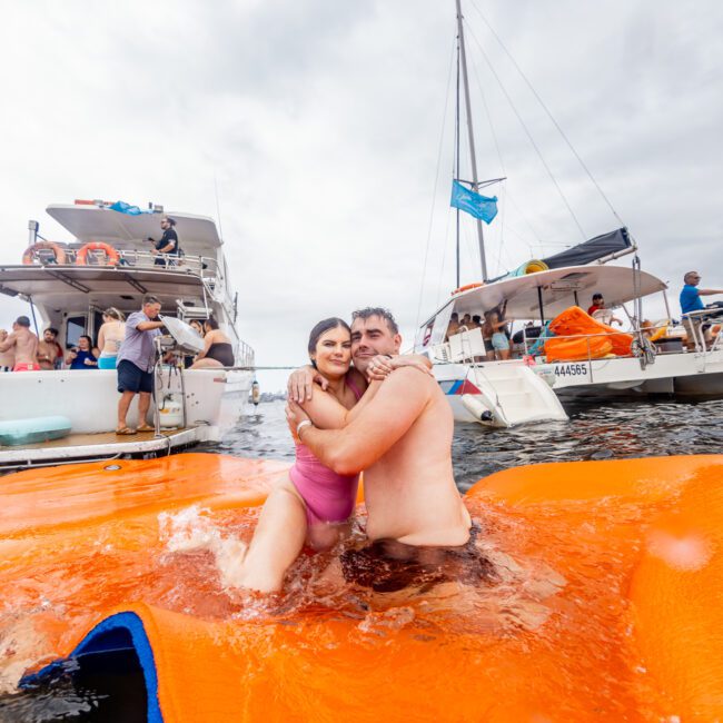 A man and a woman embrace in the water while standing on an orange float. Behind them, boats with people onboard are visible, with some individuals lounging and others engaging in various activities. The vibrant scene is part of The Yacht Social Club Event under a cloudy sky.