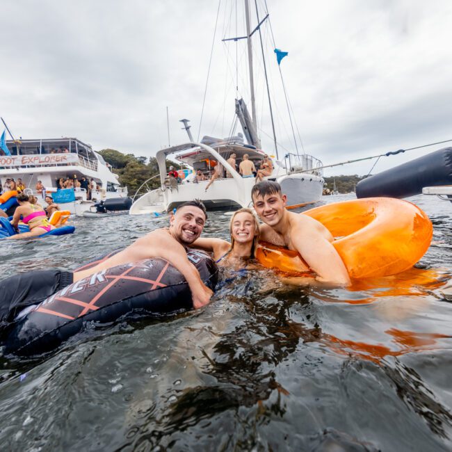 Three people smiling and holding onto an orange inflatable ring in the water, surrounded by boats. In the background, The Yacht Social Club party boat full of people dressed in blue and orange creates a lively atmosphere. The sky is cloudy, adding to the vibrant scene at Sydney Harbour Boat Hire.