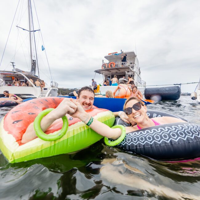 A group of smiling people having fun in the water near boats. Two people in the foreground are lounging on inflatable tubes, with one on a watermelon float and the other on a black and white float. The sky is cloudy, and various boats from The Yacht Social Club are anchored nearby.