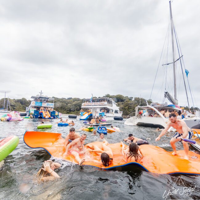 A lively summer scene of people, including children and adults, enjoying a floating orange mat on the water surrounded by boats. Others are swimming and playing nearby. Multiple boats from The Yacht Social Club are anchored close together in the background. The atmosphere is festive and fun.