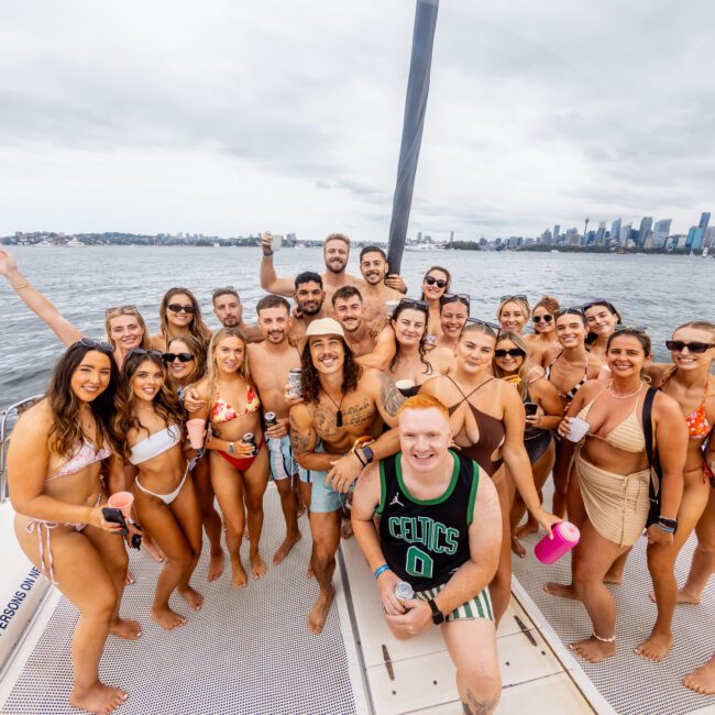 A large group of people smiling and posing for a photo on the deck of a boat. The group consists of men and women in swimwear, with some holding drinks. The background shows a city skyline and cloudy skies over a body of water, highlighting the enjoyable experience with Luxury Yacht Rentals Sydney.