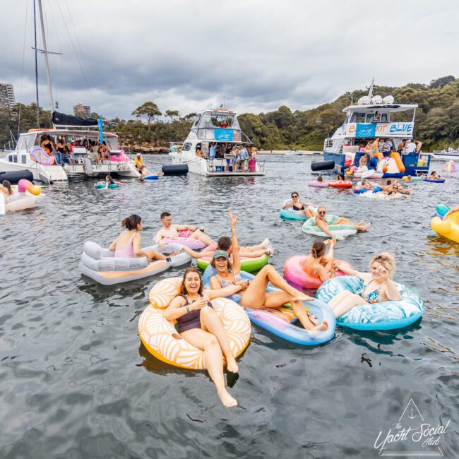 A group of people relaxing on large inflatable floats on a lake. They are smiling, wearing swimwear, and appear to be enjoying a social gathering. Several boats from The Yacht Social Club are anchored nearby, with more people visible on them, all surrounded by trees and greenery under a cloudy sky.