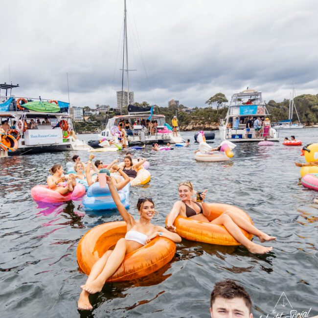 A group of people on inflatable tubes relax and have fun in a body of water, with yachts anchored in the background. The group is smiling and enjoying summer weather, surrounded by colorful inflatables and picturesque scenery. Experience this at The Yacht Social Club with Luxury Yacht Rentals Sydney.