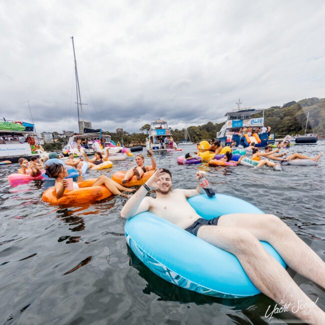 A crowd of people is seen floating in the water on various inflatable rafts. The foreground features a man on a blue raft, raising one hand in a wave and holding a drink in the other. Boats and cloudy skies are visible in the background, capturing the vibrant atmosphere of The Yacht Social Club Event.