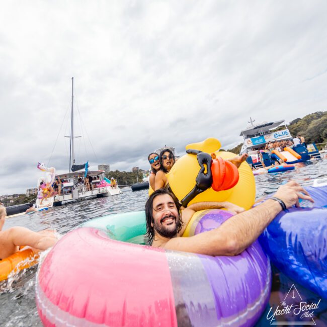 A group of people enjoy floating on colorful inflatables in the water near a boat party. The main focus is on a man smiling at the camera, leaning on a vibrant inflatable ring. Other revelers and boats from Boat Parties Sydney The Yacht Social Club are visible in the background under a cloudy sky.