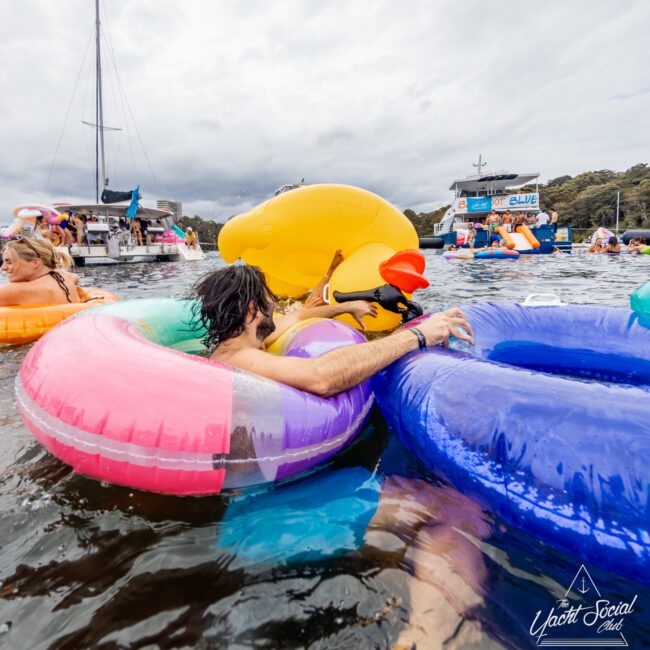 People relaxing on colorful inflatable floaties, including a large yellow duck, in a body of water. Boats and more floaties are seen in the background. The sky is overcast. A logo in the bottom right corner reads "The Yacht Social Club Sydney Boat Hire.