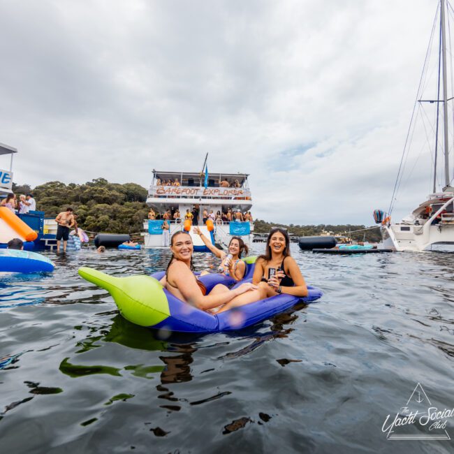 Three people are relaxing on an inflatable banana float in the water. In the background, a party on the "Barefoot Explorer" boat features several people and an Australian flag. The setting is a lively social event by the water, reminiscent of Boat Parties Sydney The Yacht Social Club events.