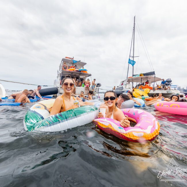 Two women are smiling and enjoying themselves while floating in the water on inflatable pool toys; one is on a turtle-shaped float, and the other is on a donut-shaped float. Behind them, more people are in the water or on yachts from Sydney Harbour Boat Hire The Yacht Social Club, creating a lively and festive atmosphere.