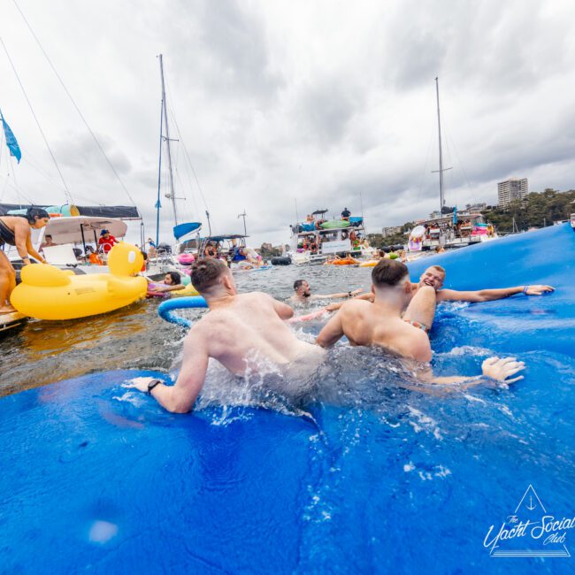 A group of people enjoying a lively time on a large floating mat in the water, surrounded by boats and inflatables. The scene is festive with cloudy skies overhead. Branding that reads "The Yacht Social Club Sydney Boat Hire" is visible in the corner of the image.