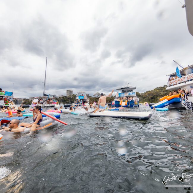 Several boats are clustered together on a cloudy day in Sydney Harbour, with people swimming and socializing in the water. Inflatable toys and water slides create a vibrant, festive atmosphere. The scene captures the lively spirit of The Yacht Social Club boat party event.