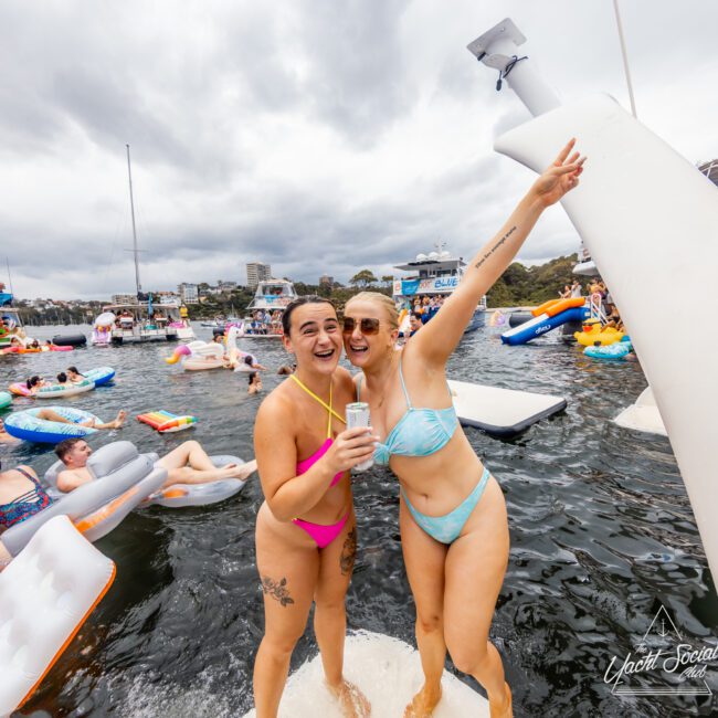 Two smiling women in swimsuits stand on a floating platform in a lake, holding drinks and posing excitedly. They are surrounded by colorful inflatables and other people in the water. The sky is overcast, and there are boats and buildings in the background, hinting at a fun gathering like The Yacht Social Club Event Boat Charters.