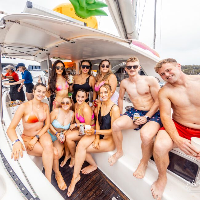 Group of nine people smiling and posing together on a boat. Seven women in bikinis and two men in swim trunks sit on the edge, enjoying drinks. Other boats and water scenery are visible in the background. A lively and festive atmosphere is evident, perfect for an adventure with Luxury Yacht Rentals Sydney.