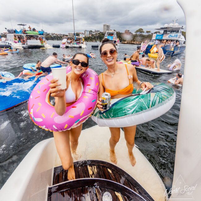 Two women in swimsuits, wearing inflatable rings (one pink donut and one green leaf), stand on a boat's platform, holding drinks. They are surrounded by people on floats and boats in the water under a cloudy sky. The Yacht Social Club Sydney Boat Hire exudes a festive summer vibe.