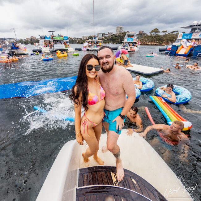 A couple in swimwear stands on a boat platform, smiling at the camera. The woman is wearing a pink bikini, and the man has swim trunks on. Other people float on inflatables in an energetic water area, with several boats in the background under a cloudy sky, embodying The Yacht Social Club's vibrant atmosphere.