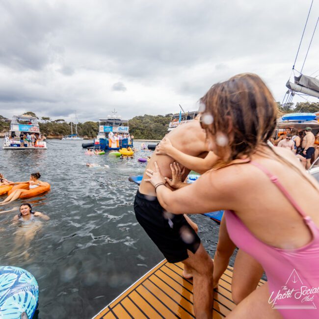 A group of people enjoy a yacht party hosted by The Yacht Social Club. A woman in a pink swimsuit playfully pushes a man in black swimwear into the water from a dock. Several others are swimming or floating on inflatables nearby. Boats and lush greenery are visible in the background.