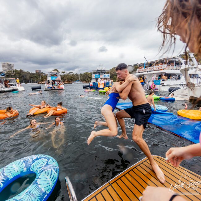 Two people, a man and a woman in swimwear, are jumping off a boat platform into the water, holding onto each other. In the background, numerous people are enjoying the water on floats and other boats are docked. It appears to be a festive event by The Yacht Social Club Sydney Boat Hire.