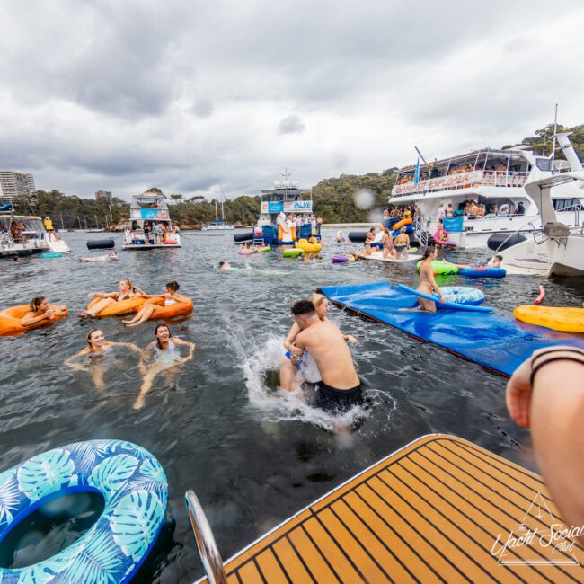 A lively scene of people enjoying a party on the water. Individuals are floating on inflatable tubes, swimming, and engaging in a playful wrestling match. Several boats, courtesy of The Yacht Social Club Sydney Boat Hire, are docked nearby with more people socializing on board. The sky is cloudy.