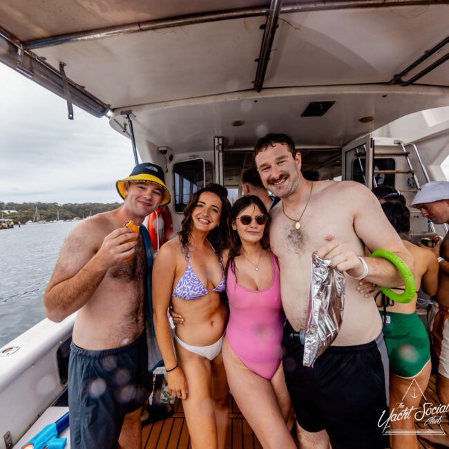 A group of four people in swimwear stand together smiling on a boat. The man on the left in a yellow hat holds a snack, while the others in bikinis and swim trunks pose closely. Enjoy unforgettable moments with Luxury Yacht Rentals Sydney at The Yacht Social Club Event Boat Charters.