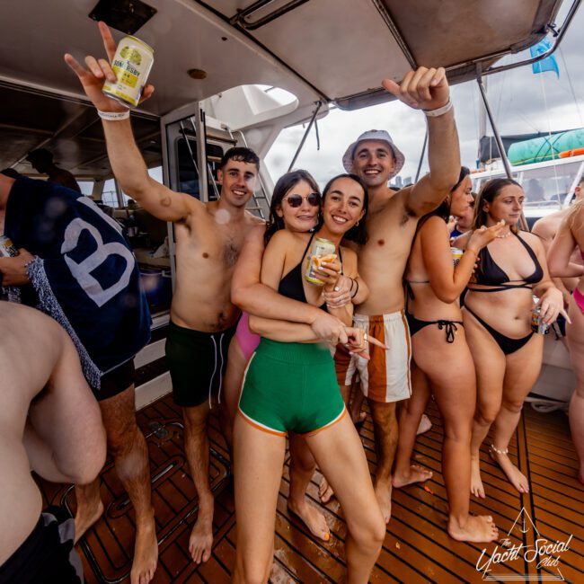 A group of young adults, wearing swimsuits, are gathered on the deck of a boat. They smile and pose for the camera, holding drinks, and appear to be enjoying a festive atmosphere. The deck is crowded with other people in swimwear. The scene is set on a sunny day during The Yacht Social Club event hosted by Sydney Harbour Boat Hire.