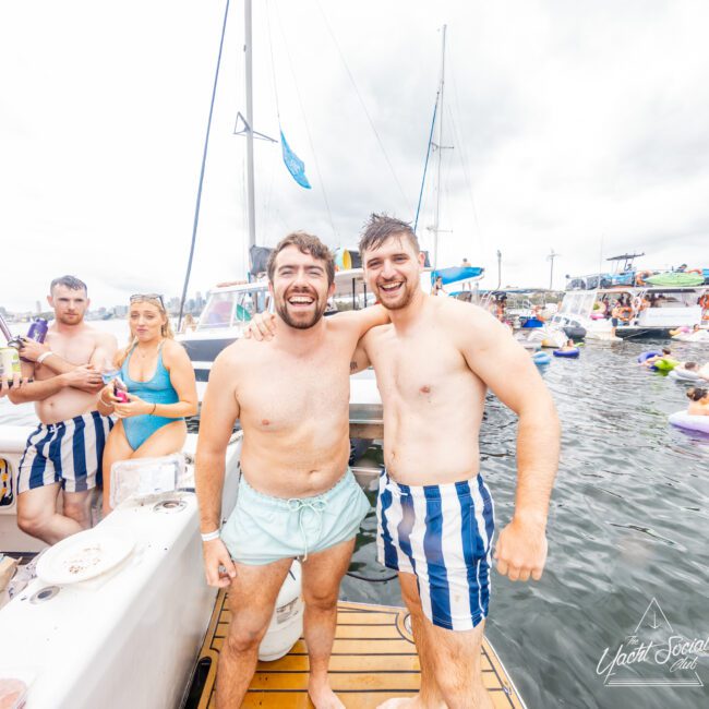 A group of friends enjoys a boat party with The Yacht Social Club. Two men in swim trunks, one in light green and the other in blue and white stripes, stand smiling at the camera. The others relax on and around the luxury yacht, with other boats and a cloudy sky in the background.