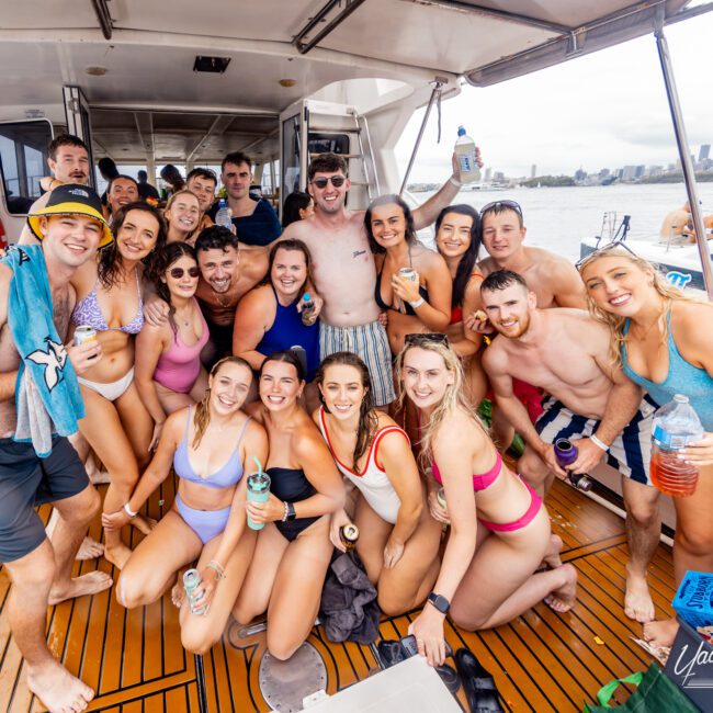 A large group of young adults wearing swimwear, smiling and posing together on a boat deck by The Yacht Social Club Event Boat Charters. Some hold drinks, others wear sunglasses or hats. The backdrop shows calm water and a distant city skyline under a cloudy sky. The mood is lively and celebratory.