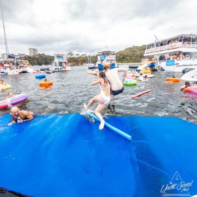 Two people jump into the water from a floating mat, surrounded by others swimming and floating on inflatables. Boats and yachts anchored in the background enhance the fun atmosphere under a cloudy sky. The Yacht Social Club logo is visible in the bottom right corner, showcasing their Sydney Harbour Boat Hire services.