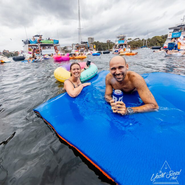 A man and a woman enjoying a day in the water, floating on a blue mat. The man is holding a can, and both are smiling. The scene is lively with numerous colorful inflatables and boats behind them under an overcast sky. The Yacht Social Club Sydney Boat Hire logo is seen at the bottom right.