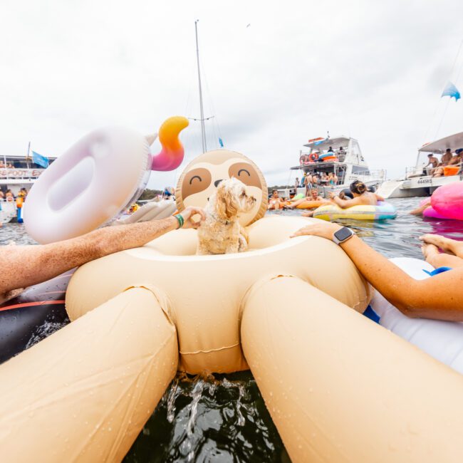 People are enjoying a day on the water, surrounded by colorful inflatable floaties, including a sloth and a donut. In the background, several boats from The Yacht Social Club can be seen. The atmosphere is lively and festive.