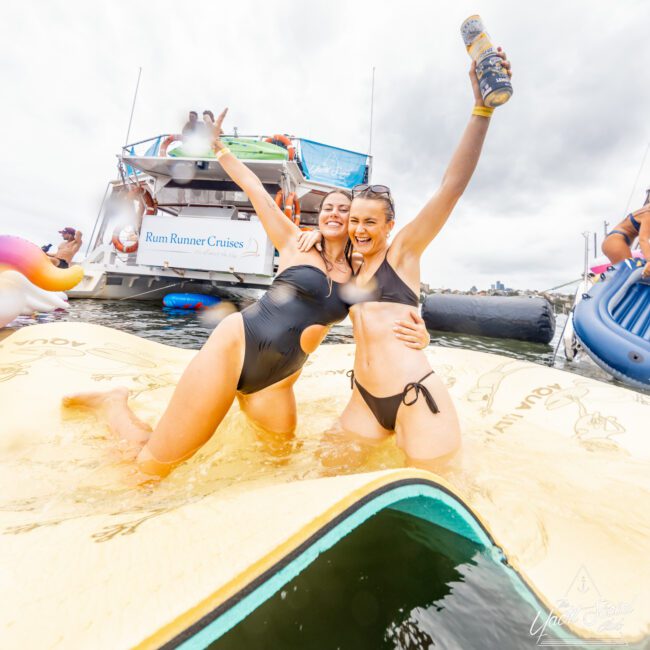 Two women in black swimsuits cheerfully pose on an inflatable pad in the water. One holds a can, and they're both smiling with arms raised. Behind them, a boat named "Rum Runner Cruises" and other flotation devices are visible. The Yacht Social Club Event Boat Charters set a festive and lively atmosphere.