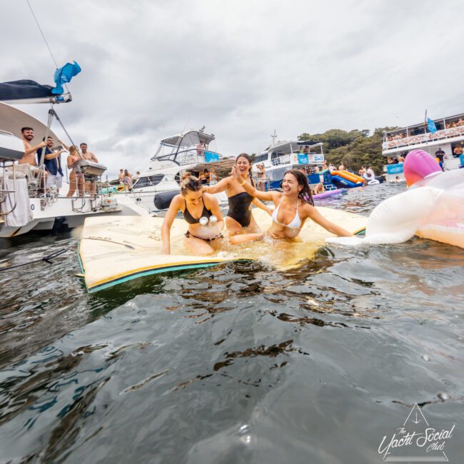 Three women in swimwear are having fun on a floating platform in the water, with anchored yachts and other people in the background. They are laughing and splashing, surrounded by an inflatable unicorn. The scene appears lively and joyful—a perfect day for Luxury Yacht Rentals Sydney or Boat Parties with The Yacht Social Club.