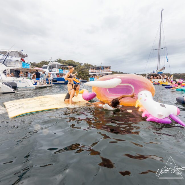 People enjoying The Yacht Social Club's event on the water, with multiple floaties, including a unicorn. Other boats are visible in the background. The atmosphere is festive, with people swimming and relaxing on inflatables. The sky is overcast. Iconic logo in the corner.