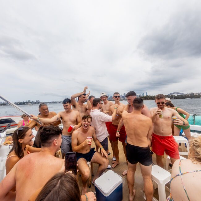 A lively group, mostly shirtless men in swimwear, are celebrating on a boat. Some hold drinks and smile while enjoying The Yacht Social Club Sydney Boat Hire. The cloudy sky serves as a backdrop to the cityscape and water.