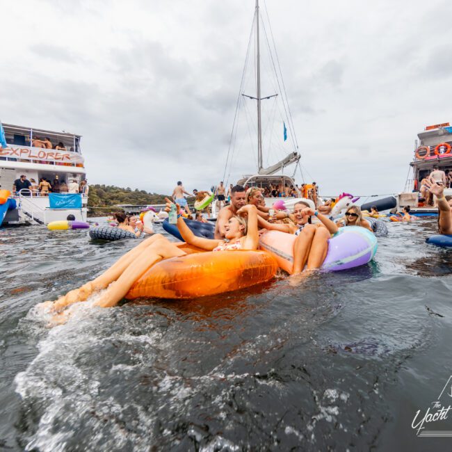 People are enjoying a party on the water, using various inflatables. There are two large boats in the background with others sunbathing, chatting, and relaxing. The event by Luxury Yacht Rentals Sydney seems festive with participants lounging in the water under a cloudy sky.