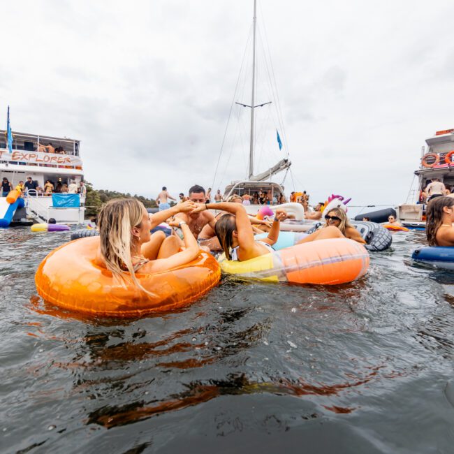 People are lounging on colorful inflatable floats in the water near two anchored boats. The atmosphere is festive with people enjoying activities and the event. Banners on the boats promote The Yacht Social Club Sydney Boat Hire, indicating a lively boat party hosted by Boat Parties Sydney.