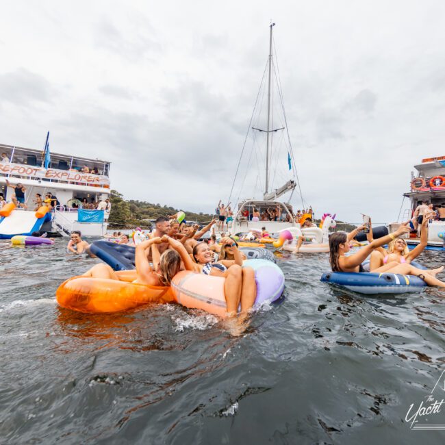 People are floating on colorful inflatable loungers in the water, surrounded by yachts and boats. The atmosphere is lively with many enjoying the social gathering, some taking photos and others lounging. Grey clouds are visible in the sky, adding a dramatic backdrop to this Yacht Social Club event in Sydney Harbour.