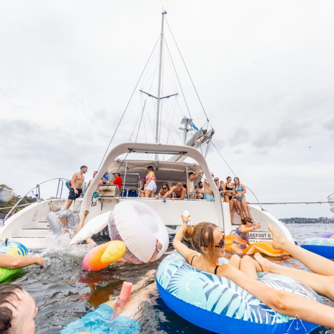 A lively group enjoys The Yacht Social Club Event Boat Charters on the water. Some guests are on the yacht while others float on colorful inflatables. The atmosphere is festive, with drinks and inflatable toys enhancing the fun under a slightly cloudy sky.