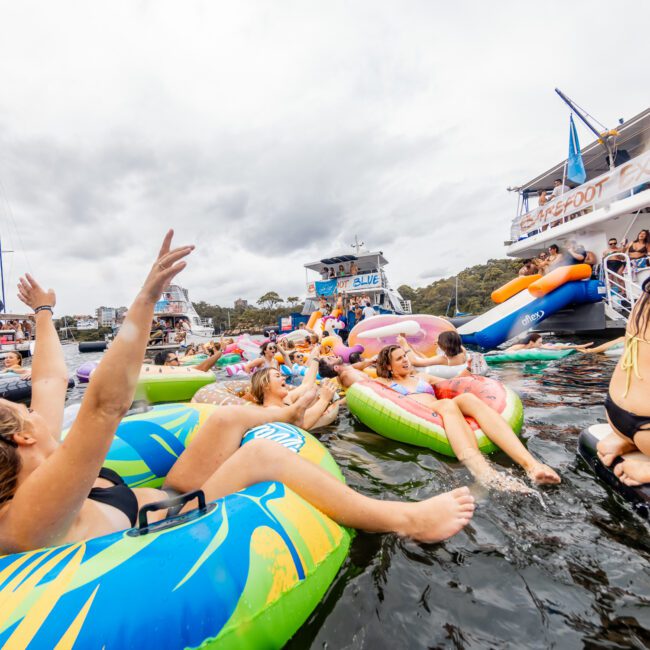 A lively scene of people relaxing on colorful inflatable tubes in the water, next to docked boats under a cloudy sky. Some are chatting, while others are enjoying the water. One person has their arms raised, and the backdrop features trees and distant buildings—a perfect day with The Yacht Social Club.