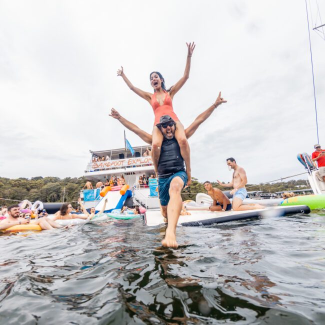 A man wades through the water with a woman on his shoulders, both smiling joyfully with arms raised. Surrounded by others in the water and on boats from The Yacht Social Club Sydney Boat Hire, they enjoy a lively outdoor gathering. Various colorful flotation devices are dotted around.