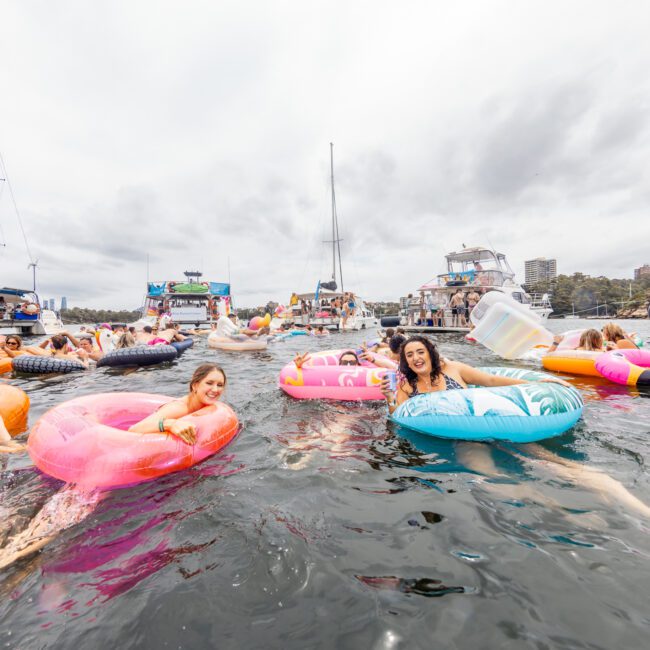 A lively scene of people enjoying a day out on the water, some holding drinks and floating on colorful inflatable rings. Boats are anchored in the background, under a cloudy sky. The atmosphere is festive and relaxed, reminiscent of The Yacht Social Club Event Boat Charters.