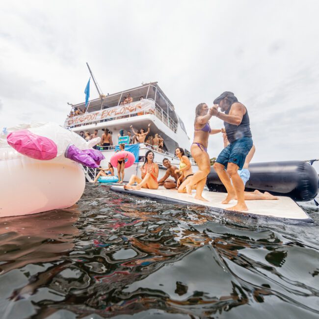 Group of people enjoying a lively party on a boat named "Party Explorer." Some float in the water, while others dance on a floating platform. There are several colorful inflatable toys, and the atmosphere is festive under a cloudy sky. The logo reads "The Yacht Social Club Event Boat Charters.