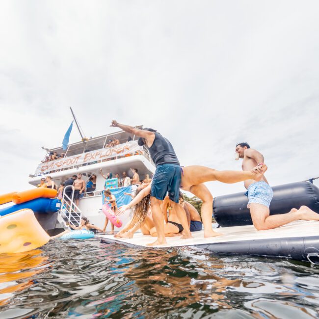 A group of people enjoying a Luxury Yacht Rentals Sydney party, with some standing on an inflatable pad at the back of a yacht. The background shows a partially cloudy sky, and the water is calm. The yacht is decorated with banners and inflatables, and there's a social atmosphere. "Yacht Social Club" is written on the image.