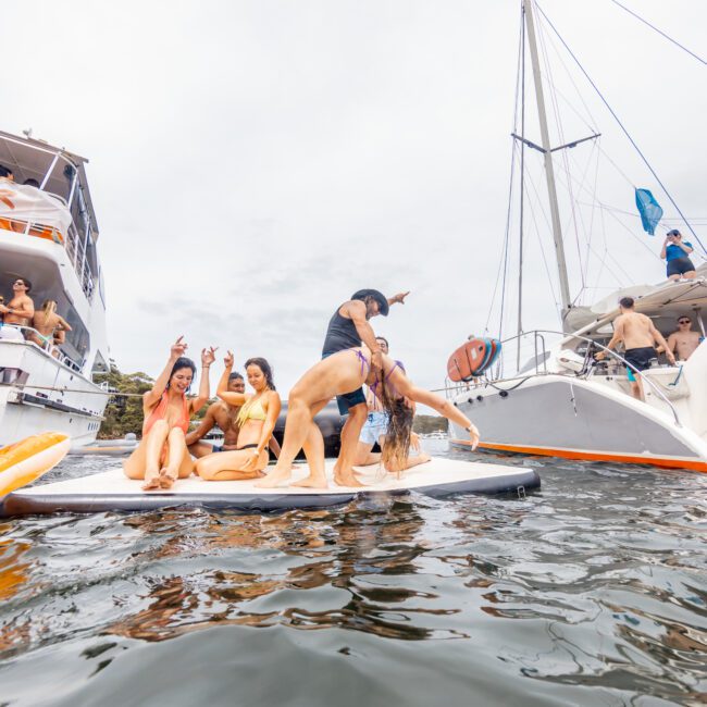 A group of people are having fun on a boat and paddleboard in a lively outdoor water setting. Some are waving and smiling at the camera, while one person playfully bends over another on the paddleboard. Several boats from The Yacht Social Club Event Boat Charters are anchored nearby, reflecting their activities in the water.