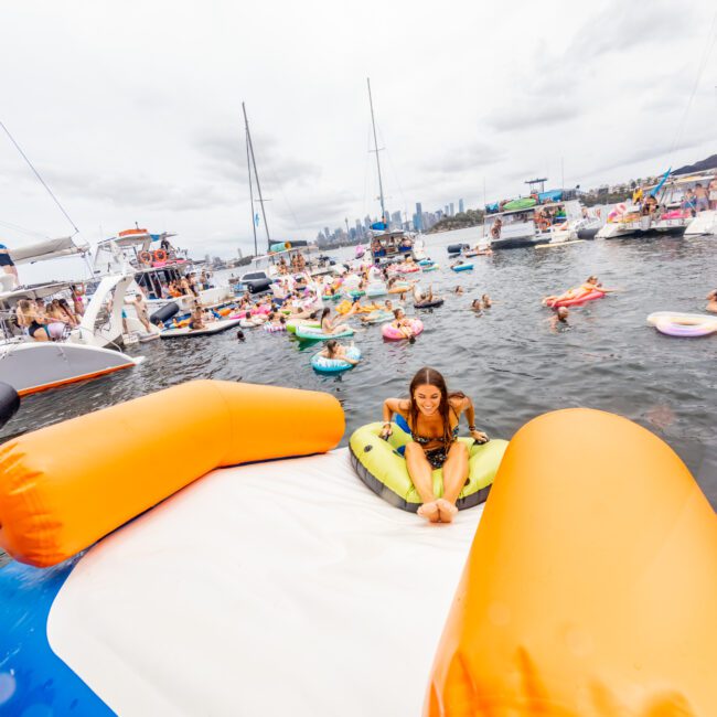 A lively scene on a lake features various boats and people on inflatable rafts. In the foreground, a woman in a swimsuit sits on an orange and white inflatable structure. The skyline of a city is visible in the background under a cloudy sky. Experience it all with Sydney Harbour Boat Hire The Yacht Social Club.