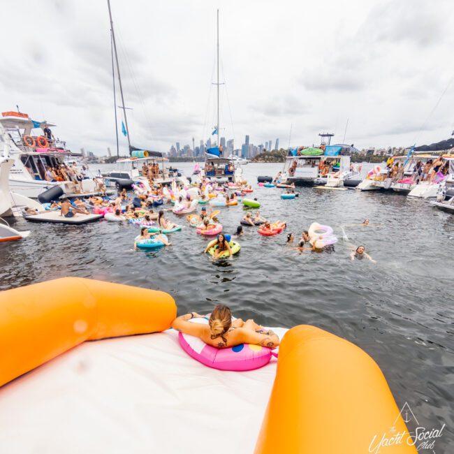 A lively crowd enjoys a yacht party on a cloudy day, hosted by The Yacht Social Club Sydney Boat Hire. People float on colorful inflatables and swim between several boats docked closely together. The city skyline is visible in the background, with an inflatable water slide in the foreground.