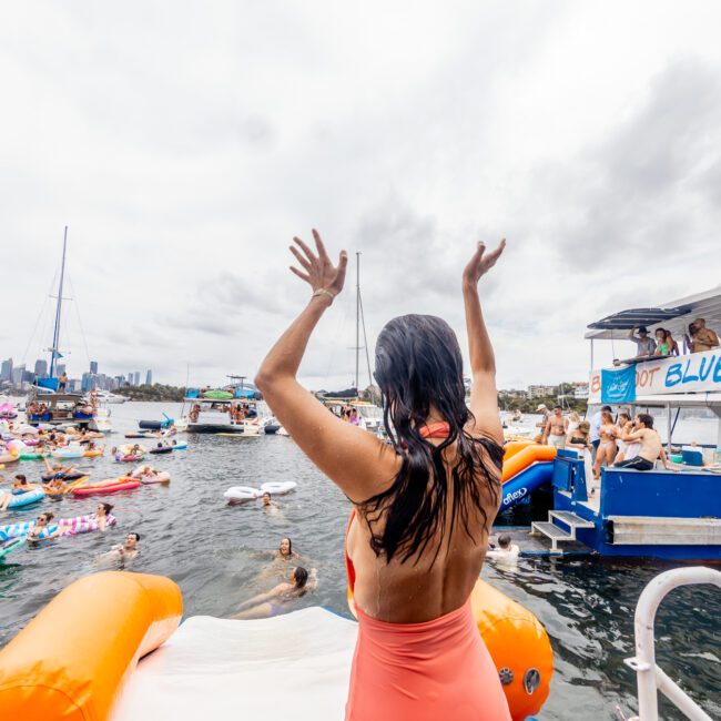 A person stands with arms raised on an inflatable slide attached to a boat. The boat, named "Get Blue," is surrounded by people swimming and using various inflatables in the water. Other boats and a city skyline are visible in the background, creating the perfect scene for The Yacht Social Club Sydney Boat Hire.