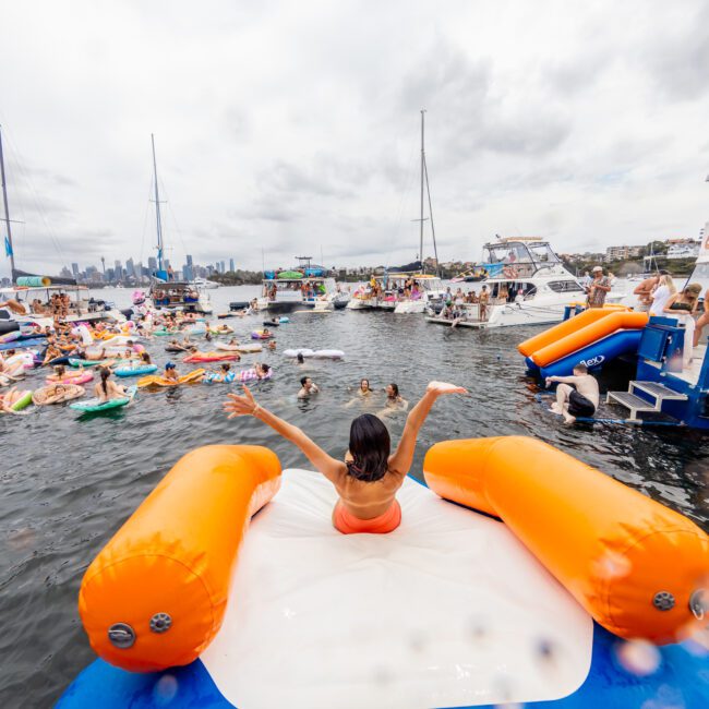 A crowded boat party on a lake with many people enjoying the water on colorful inflatables and boats. The focus is on a person sitting on a large orange and white inflatable with arms raised, facing away from the camera. The weather is cloudy, and the shore is visible in the background. It's reminiscent of a fun day at The Yacht Social Club Event Boat Charters.