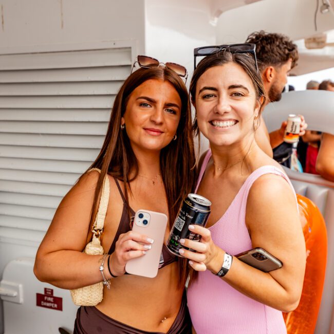 Two young women pose and smile at the camera on a boat. The woman on the left has long brown hair, wears a brown skirt and beige crop top, holding a smartphone. The woman on the right has dark hair in a pink swimsuit, holding a can. Others are in the background enjoying Boat Parties Sydney The Yacht Social Club.