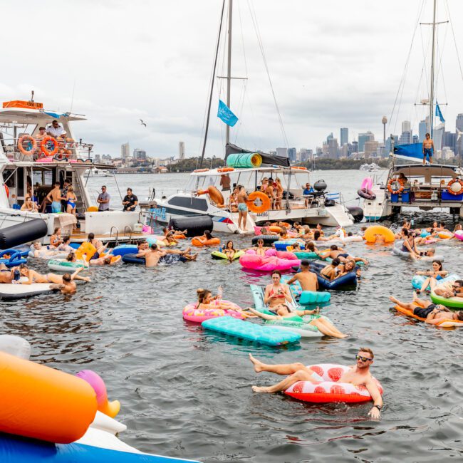 People are enjoying a festive gathering on the water, floating on colorful inflatables near docked boats. The background features a city skyline under a cloudy sky, creating a vibrant and relaxed atmosphere. It's an iconic scene often seen with Sydney Harbour Boat Hire The Yacht Social Club events.