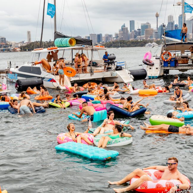 A lively scene of people on colorful inflatable floats in the water near several anchored boats. Some are relaxing, others engaging in conversation or taking photos. The skyline of a city is visible in the background under a cloudy sky, creating the perfect setting for Luxury Yacht Rentals Sydney.