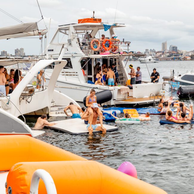 A lively scene on the water shows people enjoying a boat party. Multiple boats are docked together, with guests lounging on floats and swimming. The backdrop features a cloudy sky and a distant city skyline, creating a festive and relaxed atmosphere. Experience this with Luxury Yacht Rentals Sydney from The Yacht Social Club.